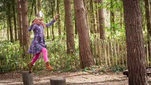 A child leaps across tree stumps in the natural play area at Calke Abbey, surrounded by green woodland and wearing Easter bunny ears.